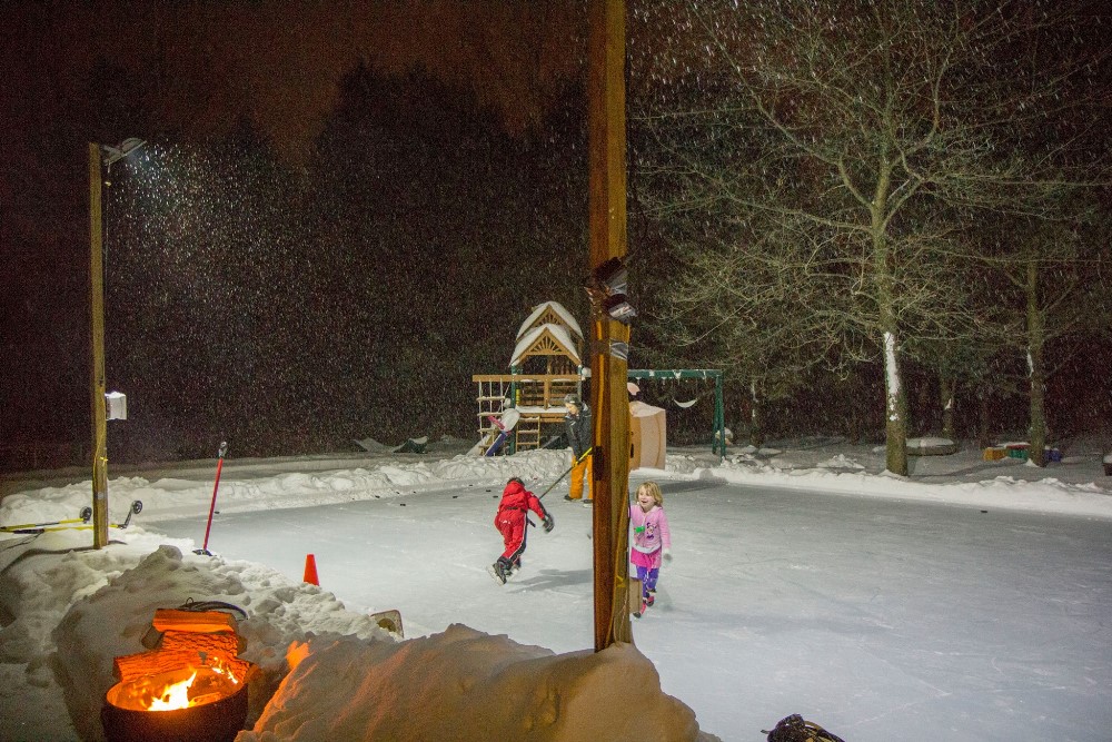 Kids skating under the lights and snow on a backyard rink.